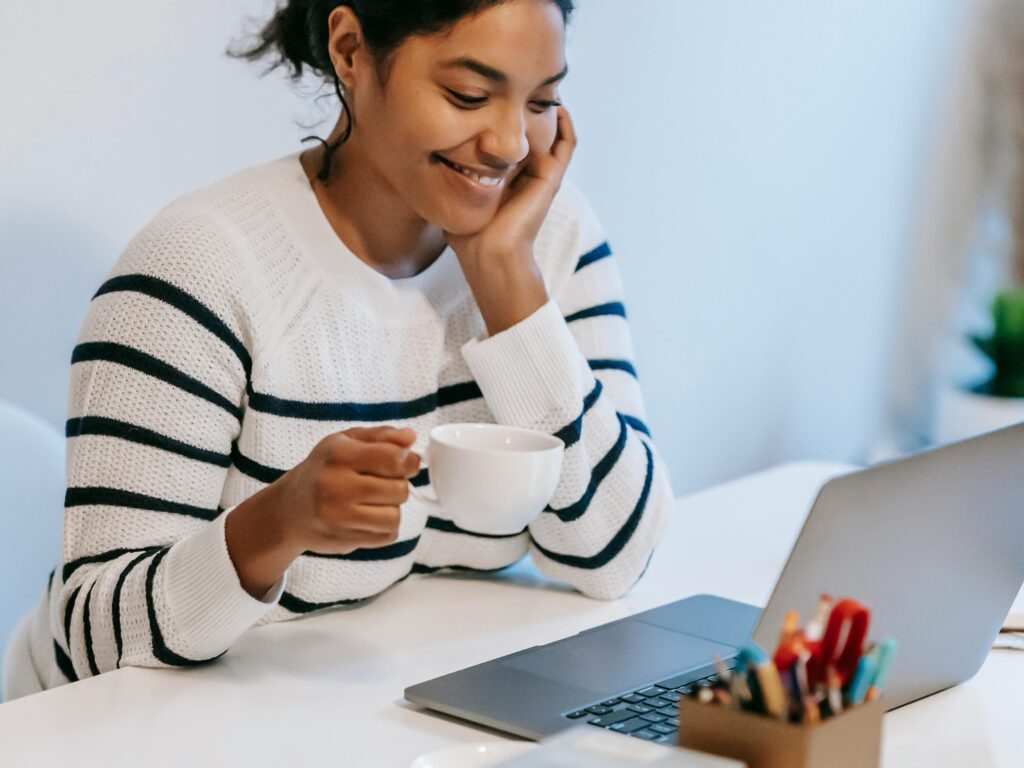 Positive young African American female freelancer in casual clothes talking on smartphone and drinking coffee while working remotely on laptop at home